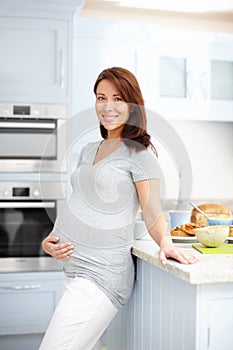 Catering for two. Portrait of a beautiful pregnant woman standing in the kitchen holding her stomach.