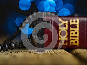 Catholic cross on a rosary and a book - the Holy Bible on a wooden table. Dark blue background with twinkling lights. Faith,