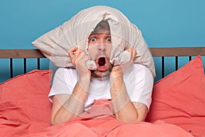 Cauacsian young man with pillow on head lying in bed and screaming