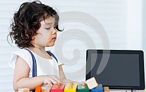 Caucasian adorable cute curly hair little girl wearing white shirt, staying alone, playing toys and tablet in living room at home