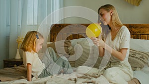 Caucasian adult mother with daughter sitting playing on bed in bedroom mum blowing yellow balloon blow air in inflatable