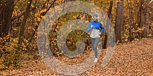 Caucasian adult woman in headset and blue sportswear is jogging in the forest in golden autumn, format image 1x2