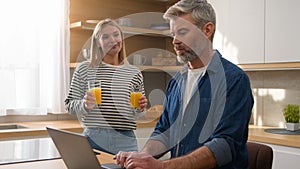 Caucasian adult woman pour orange juice give glass husband man working online with laptop in home kitchen clinking