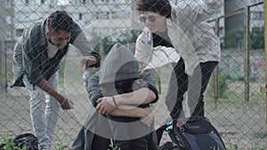 Caucasian and African American 1990s-styled men bullying young curly-haired guy sitting at mesh fence. Hooligans making