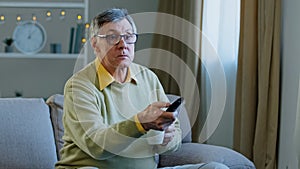 Caucasian aged senior mature man in glasses relax on couch at home change channel with remote controller watching TV