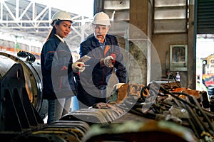 Caucasian and Asian factory worker discuss together in front of stack and layer of metal part of machine that use for industrial