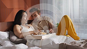 Caucasian and asian men sitting on bed in cozy apartment and enjoying breakfast