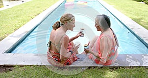 Caucasian and Asian senior women enjoy drinks by the pool, clad in swimwear