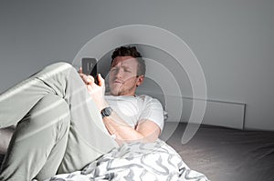 Caucasian attractive young man in white t-shirt woke up chatting, surfing, working on pc and making phone call on bed in bedroom