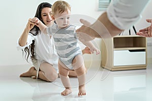 Caucasian baby boy child learn to walk with parents support in house. Happy family, mother and father helping young toddler son