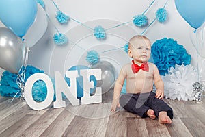 Caucasian baby boy in dark pants and blue bow tie celebrating his first birthday with letters one and balloons