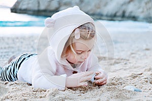 Caucasian baby girl wearing shirt with funny animal ears on sandy beach playing with stones,eating sand by blue sea