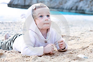 Caucasian baby girl wearing shirt with funny animal ears on sandy beach playing with stones,eating sand by blue sea