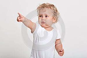 Caucasian baby pointing with index finger away, sees something interesting, wearing t shirt, standing against white wall, charming