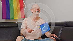 Caucasian bald gay man waving European flag, sitting on a sofa, smiling, and holding remote control.