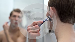 Caucasian bearded handsome man shaves with a disposable razor in the bathroom, looking in the mirror. Guy shaving his