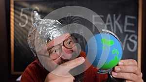 Caucasian bearded man in a foil hat on the background of a chalk board. The schizophrenic holds the globe in his hands