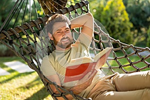 Caucasian bearded young man lying in hammock with a book in hands