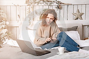 Caucasian beautiful girl sits on bed with laptop and types on the phone light christmas tree