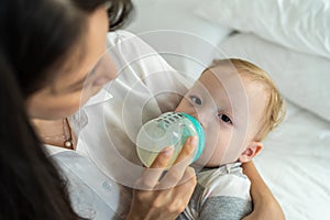 Caucasian beautiful mother holding and feeding baby from milk bottle. Attractive loving family, mom sit on bed and carry sleepy