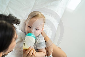 Caucasian beautiful mother holding and feeding baby from milk bottle. Attractive loving family, mom sit on bed and carry sleepy