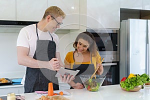 Caucasian beautiful young husband and wife searching menu recipe in digital tablet in kitchen at home. Happy love couple and