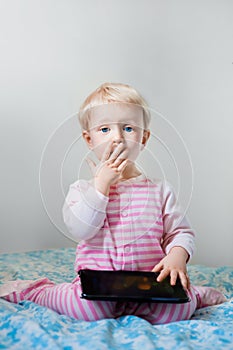 Caucasian blond baby girl sitting in bed playing with digital tablet with funny face expression