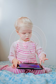 Caucasian blond baby girl sitting in bed playing with digital tablet with funny face expression