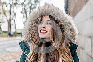 Caucasian blond girl with coat and a big hood smiles at the camera on a cold day