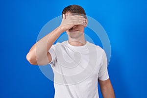 Caucasian blond man standing over blue background covering eyes with hand, looking serious and sad