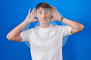 Caucasian blond man standing over blue background trying to hear both hands on ear gesture, curious for gossip