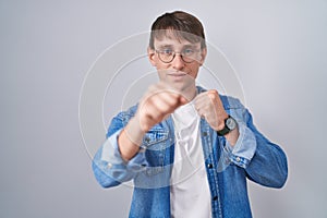 Caucasian blond man standing wearing glasses punching fist to fight, aggressive and angry attack, threat and violence