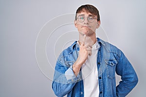 Caucasian blond man standing wearing glasses thinking concentrated about doubt with finger on chin and looking up wondering