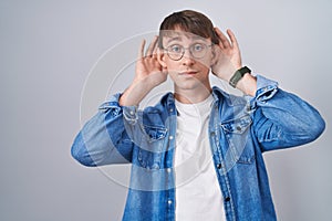 Caucasian blond man standing wearing glasses trying to hear both hands on ear gesture, curious for gossip