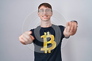 Caucasian blond man wearing bitcoin t shirt pointing to you and the camera with fingers, smiling positive and cheerful