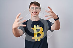 Caucasian blond man wearing bitcoin t shirt shouting frustrated with rage, hands trying to strangle, yelling mad