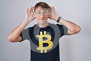 Caucasian blond man wearing bitcoin t shirt trying to hear both hands on ear gesture, curious for gossip