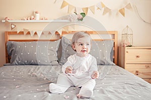 Caucasian blonde baby girl in white onesie sitting on bed in bedroom