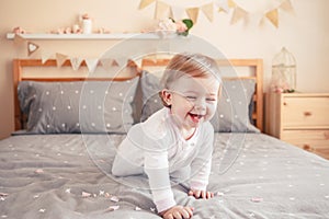 Caucasian blonde baby girl in white onesie sitting on bed in bedroom