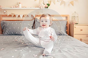 Caucasian blonde baby girl in white onesie sitting on bed in bedroom