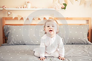 Caucasian blonde baby girl in white onesie sitting on bed in bedroom