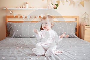 Caucasian blonde baby girl in white onesie sitting on bed in bedroom