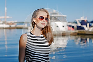 Caucasian blonde woman with tanned skin striped t-shirt and blue jeans by seashore lakeshore, with yachts boats on background