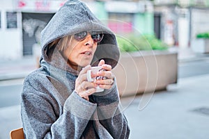 Caucasian blonde woman wearing sunglasses and coat hoodie sitting in a bar terrace having a cup of coffee