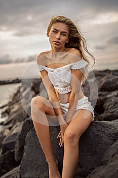 Caucasian blonde young woman relaxing on the rocky beach. Sensual girl looking at the camera, sitting on the rocks