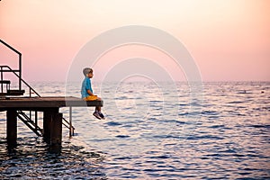 Caucasian boy in a blue striped t-shirt and yellow shorts sits on a pier, having given legs to the sea, and enjoys at sunset