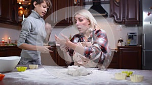 Caucasian boy bringing phone to woman kneading dough in kitchen. Busy Caucasian young mother talking on smartphone as