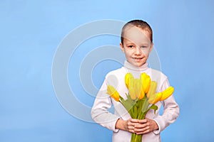Caucasian boy dressed in white jacket holds a bouquet of yellow tulip and looks at the camera. Cute little babe smiling standing