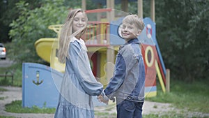 Caucasian boy and girl holding hands and turning to camera. Portrait of positive children looking at camera and smiling