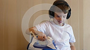 Caucasian boy listens to cheerful music with headphones, dances and does routine homework. The child ironing clothes after washing